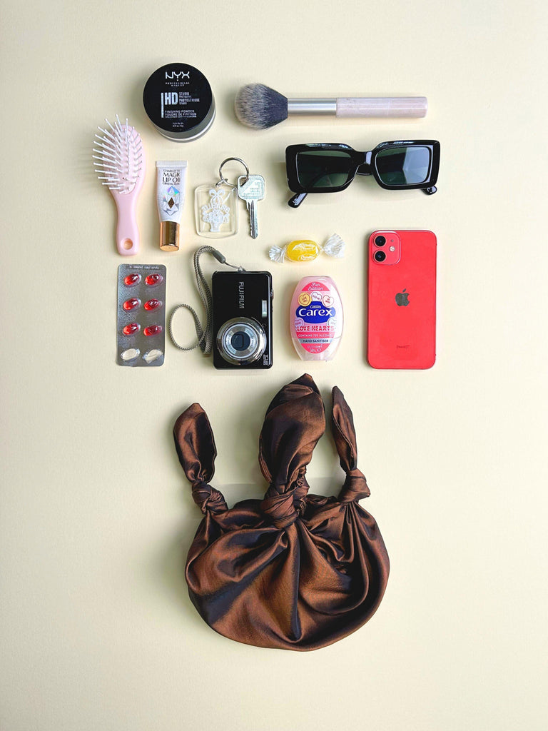 Collection of everyday items including a phone, camera, and makeup products on a beige background. A bronze taffeta handbag sits at the bottom.