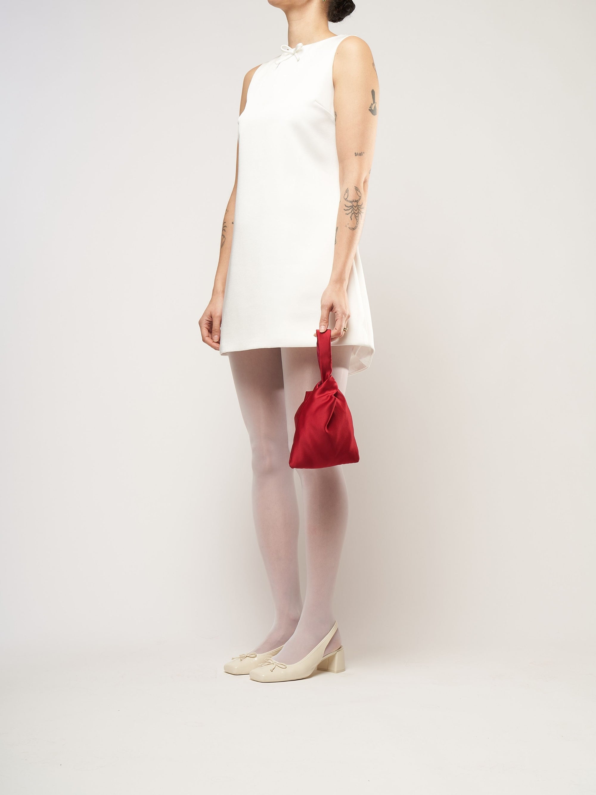 A person in a white sleeveless dress stands against a plain background. They hold a small burgundy bag and wear white tights with cream heels.