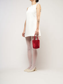 A person in a white sleeveless dress stands against a plain background. They hold a small burgundy bag and wear white tights with cream heels.