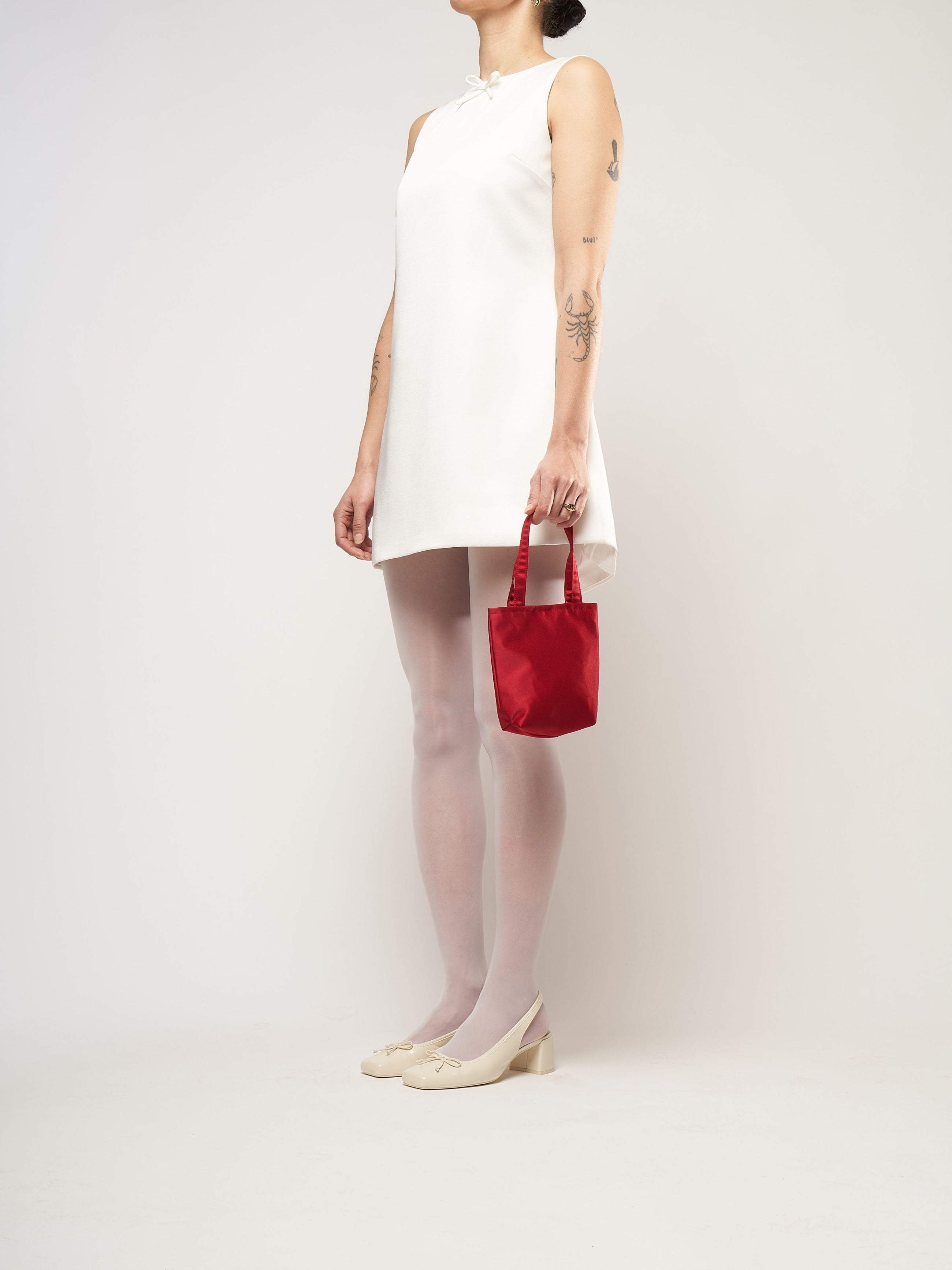 A person in a white sleeveless dress stands against a plain background. They hold a small burgundy bag and wear white tights with cream heels.