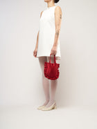 A person in a white sleeveless dress stands against a plain background. They hold a small burgundy bag and wear white tights with cream heels.