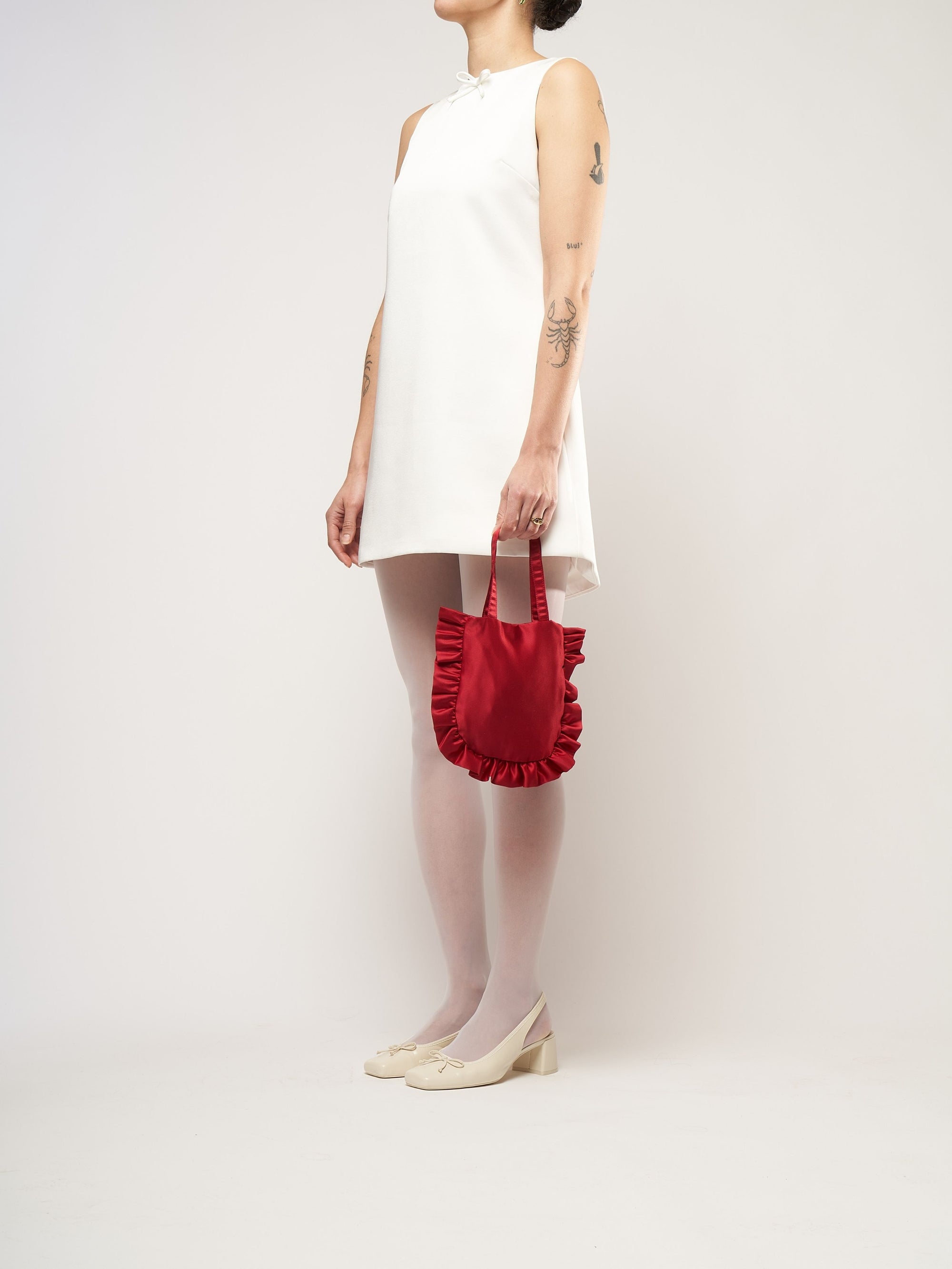 A person in a white sleeveless dress stands against a plain background. They hold a small burgundy bag and wear white tights with cream heels.