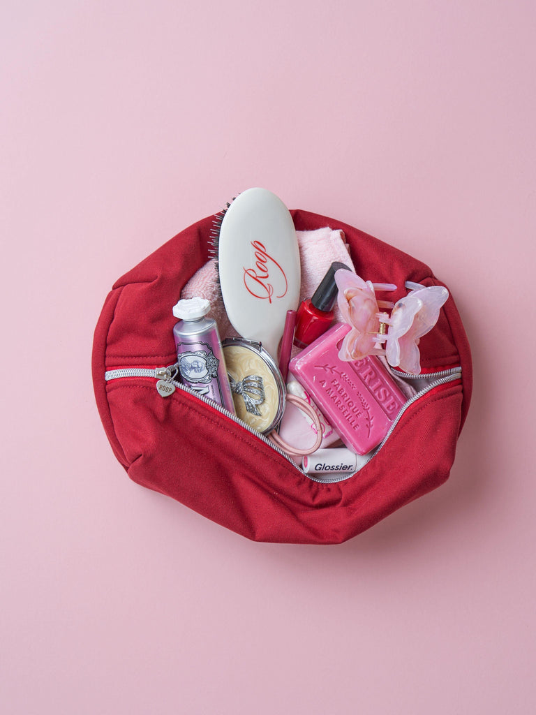 An open padded burgundy makeup and toiletry bag with various makeup products and hair accessories inside it. Set on a pink background.