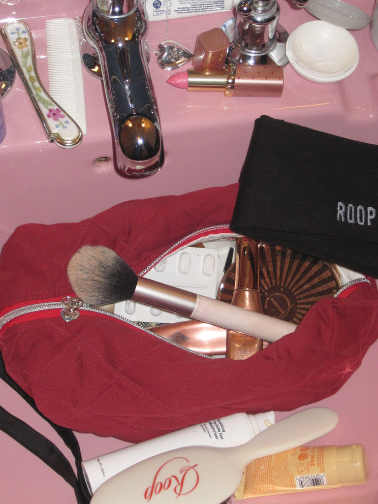 A quilted burgundy makeup bag, open revealing makeup products, sits in a pink sink with more cosmetics and hair accessories surrounding it.