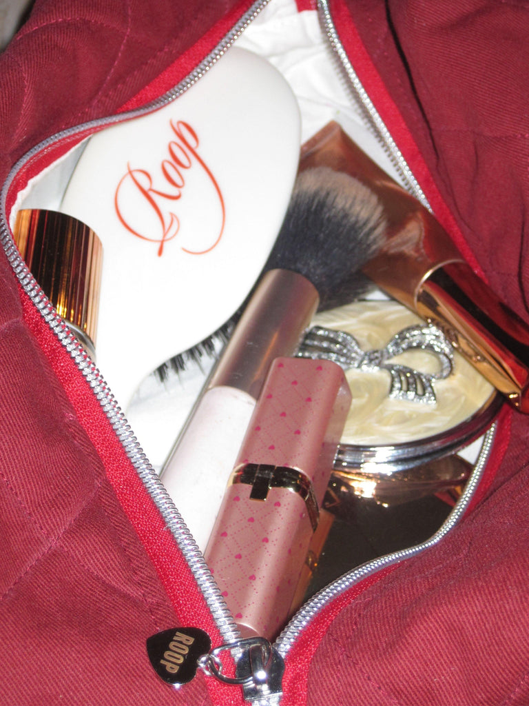 Close up of an open quilted burgundy toiletry and makeup bag. Inside are various makeup products and a white hairbrush that says ROOP on it.