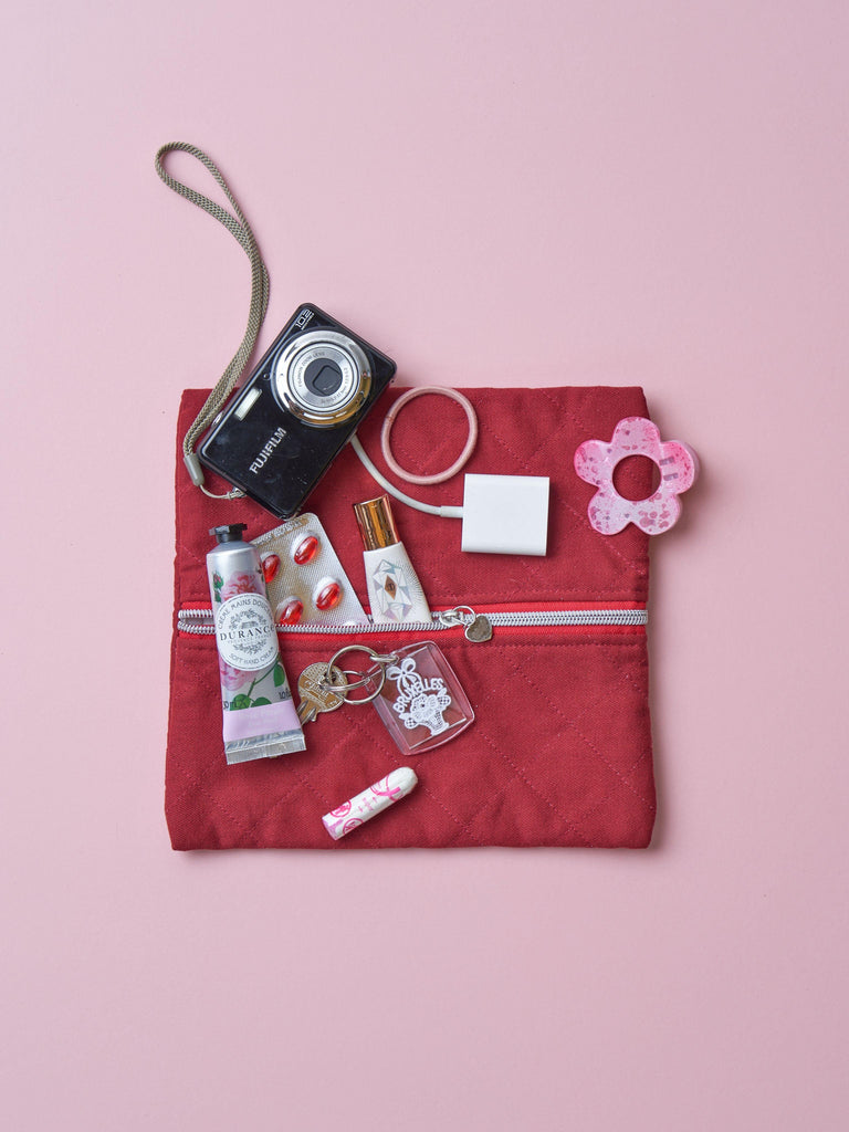 A quilted burgundy cotton zipper pouch, the zip has a heart charm on it. Various items including a camera, lip balm, and keychain are on top of it. Set on a pink background.