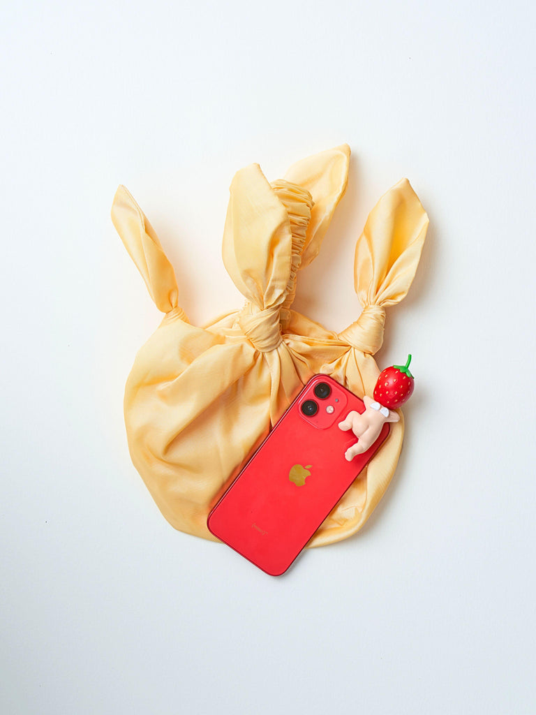 butter yellow baby furoshiki handbag with a red iphone with a strawberry sonny angel hipper lying on top of the bag. Set on a blank background.