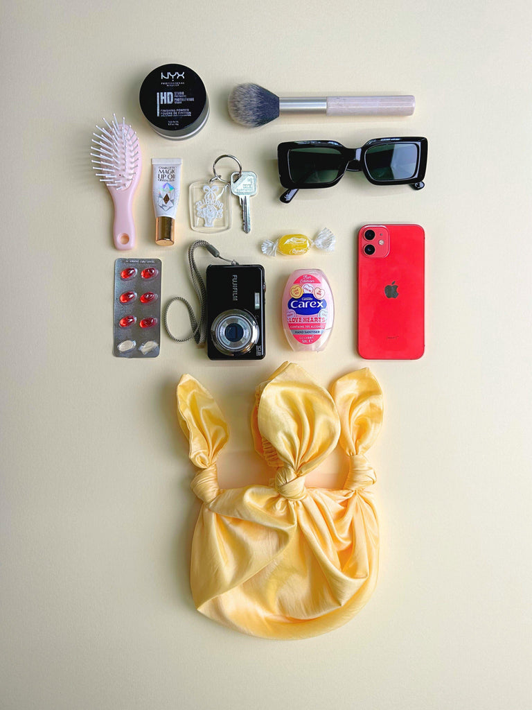 Collection of personal items including a phone, camera, and hair accessories on a light background. A yellow knotted handbag sits at the bottom.