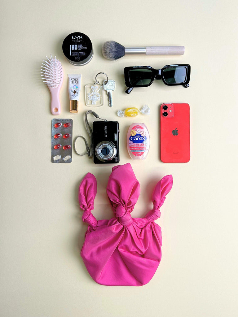 Collection of everyday items including a phone, camera, and makeup products on a beige background. A pink handbag sits at the bottom.