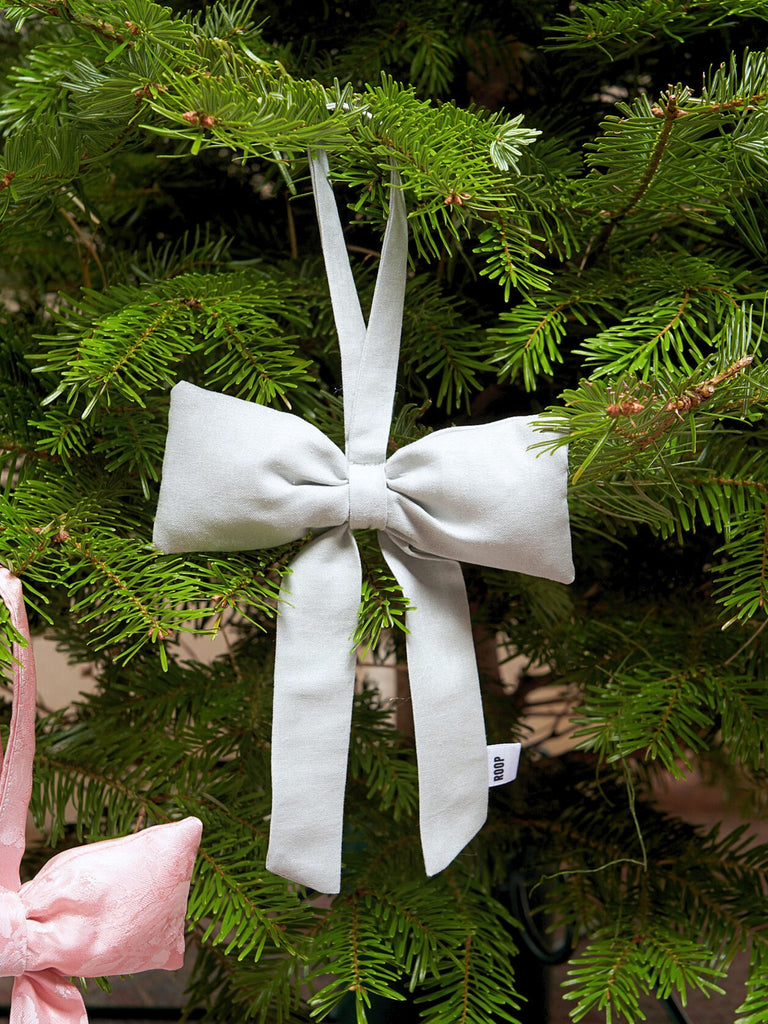 Silver / grey fabric bow with long tails hangs on a green fir tree, creating a festive and elegant contrast. A hint of a pink bow appears at the edge.