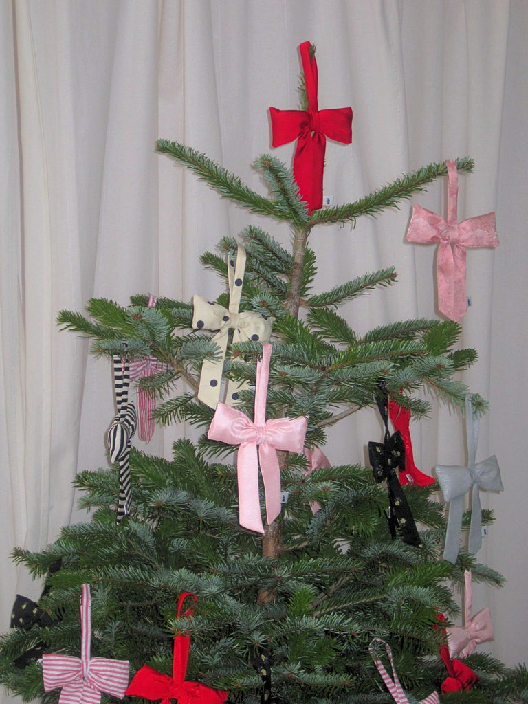 A green Christmas tree with multiple coloured fabric padded bows, including red, pink, and polka-dotted, against a light curtain background. The scene is festive and simple.