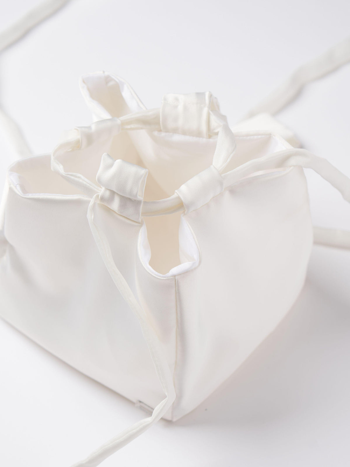 Close up of an ivory satin box drawstring handbag with ribbon drawstring details, open to reveal interior, set on a white background