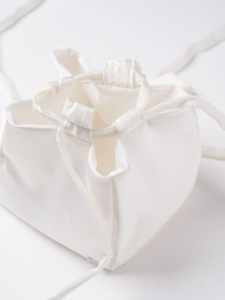 Close up of an ivory satin box drawstring handbag with ribbon drawstring details, open to reveal interior, set on a white background