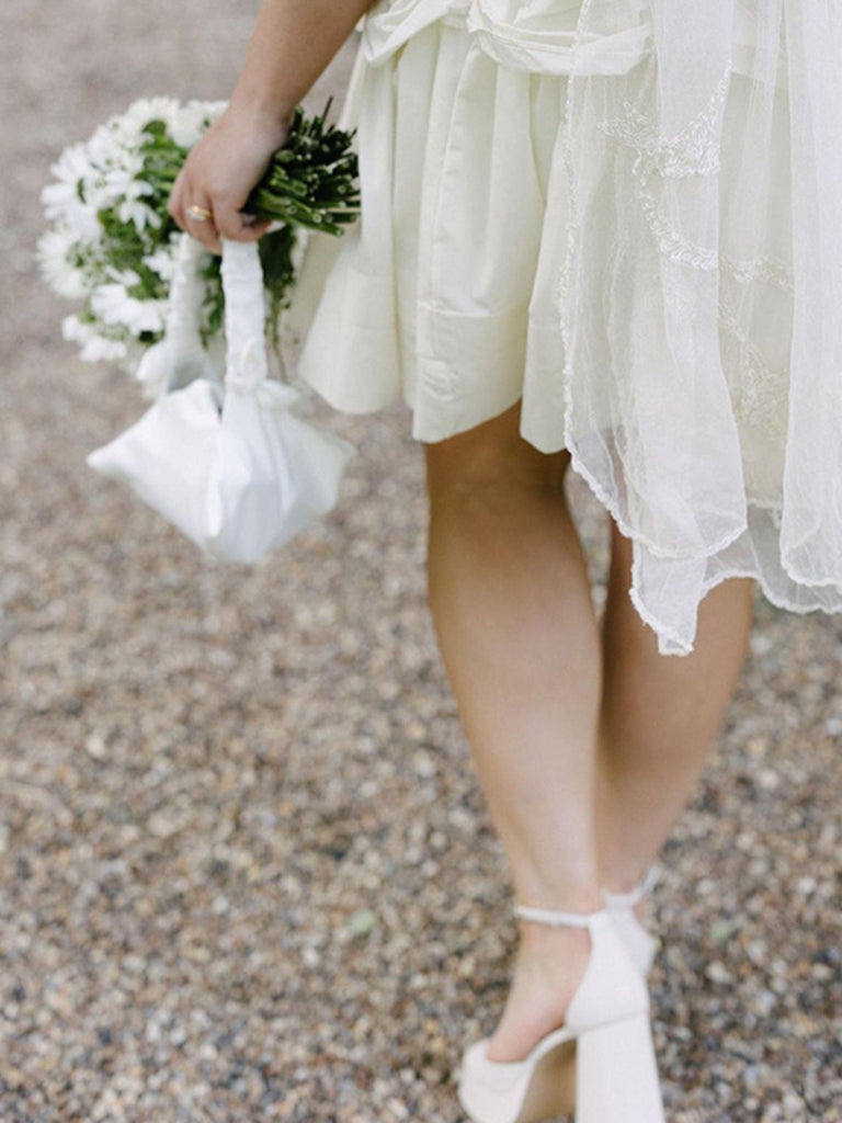 Person wearing a white dress with lace details, holding a small bouquet, and a small ivory satin ruched handle handbag, they are standing on a gravel surface.