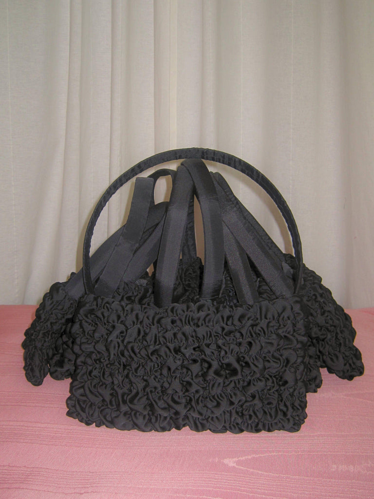 A collection of black satin textured handbags piled on top of some pink moire fabric, set against a plain background.
