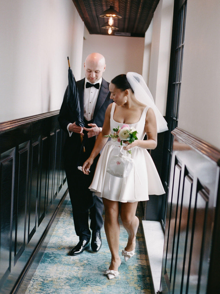 Bride and groom walking down a hallway in a formal setting. She is holding an ivory satin box bag with a red heart embroidered on it.