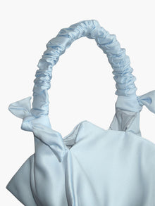 Close up of a light blue satin handbag with a ruched handle and decorative bow details, against a white background
