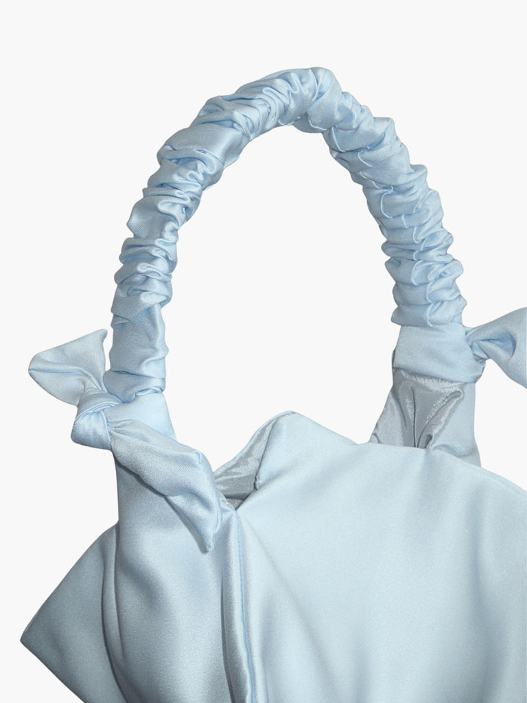 Close up of a light blue satin handbag with a ruched handle and decorative bow details, against a white background