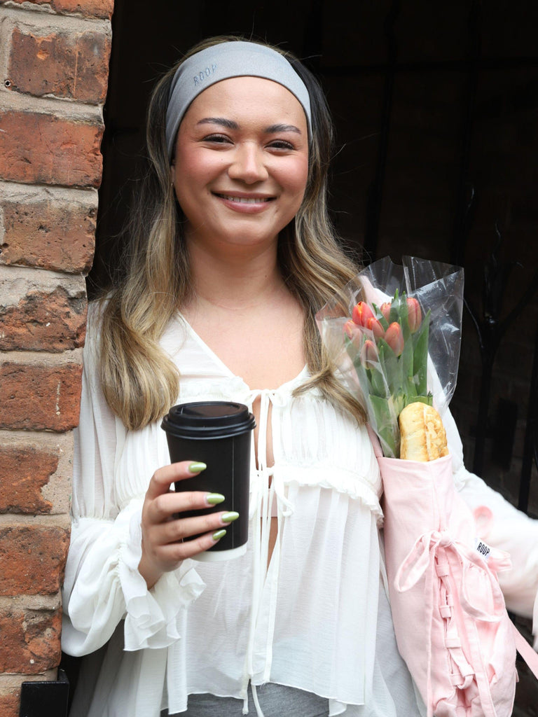 Woman holding a coffee cup and flowers against a brick wall. She wears a white flowy top and a grey stretch headband