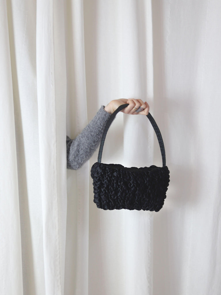A persons arm comes through a gap in some cream curtains, they are holding a black textured satin handbag.
