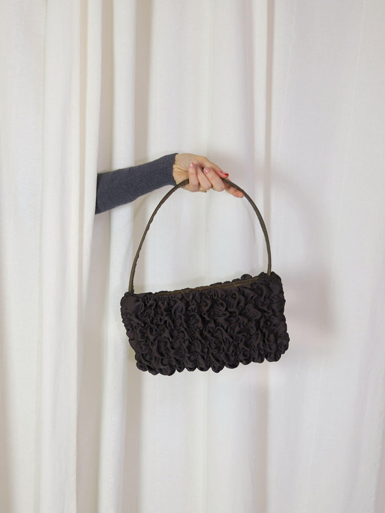 A persons arm comes through a gap in some cream curtains, they are holding a brown textured satin handbag.