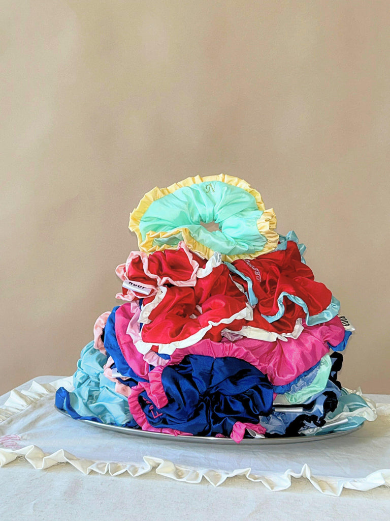 Stack of colourful ruffled taffeta scrunchies on a silver plate, sitting on a ruffled white cloth, set against a beige background.