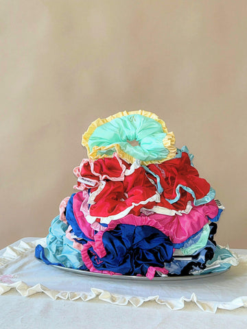 Stack of colourful ruffled taffeta scrunchies on a silver plate, sitting on a ruffled white cloth, set against a beige background.