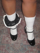 A persons legs stood on a patterned rug, they wear white socks pulled up with black patent shoes, styled with a black scrunchie with white ruffled trim and 'kim' embroidered in white