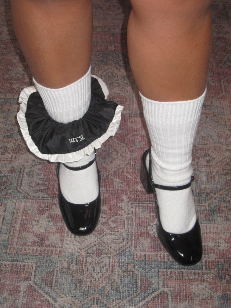 A persons legs stood on a patterned rug, they wear white socks pulled up with black patent shoes, styled with a black scrunchie with white ruffled trim and 'kim' embroidered in white