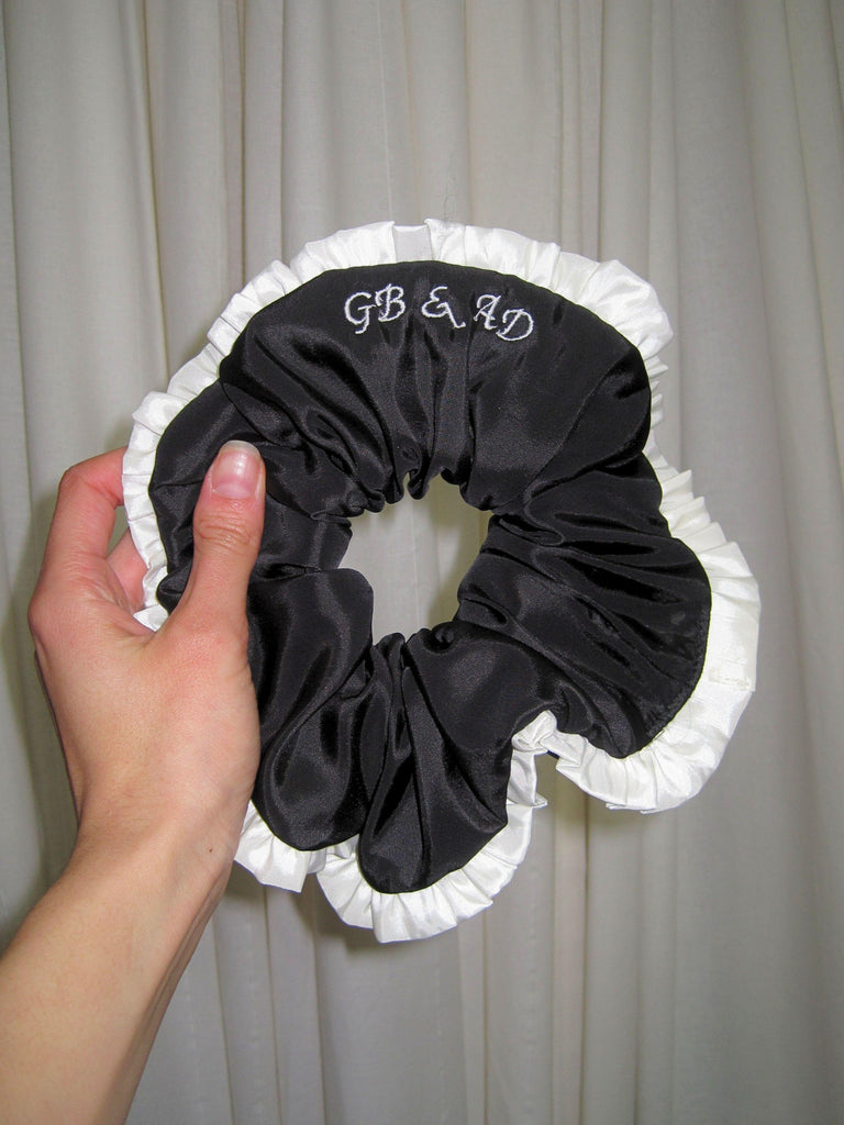 Black scrunchie with white ruffle and 'GB & AD' embroidered text held by a hand against a plain background