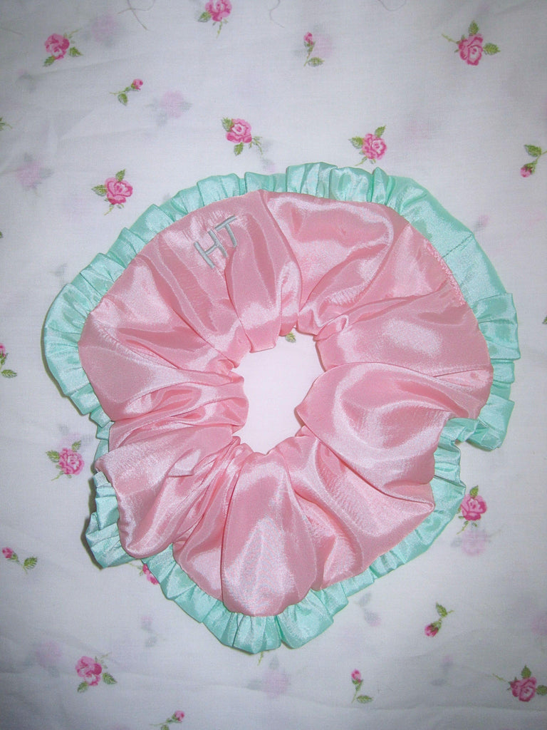 Pale pink scrunchie with mint green ruffled trim and mint green embroidered initials on a white floral background.