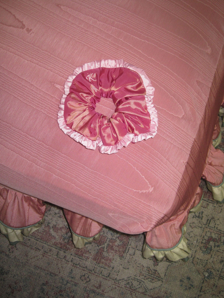 Dusky pink scrunchie with light pink ruffled edge and embroidered pink 'K' sat on a pink moire footstool on a patterned rug.