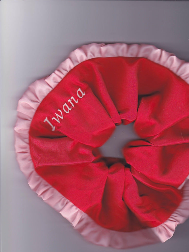 Red scrunchie with pink ruffled trim and pink embroidered name 'Iwana' on a grey background