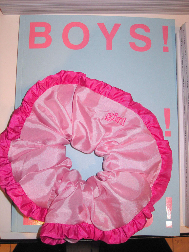 Light pink scrunchie with ruffled hot pink trim and embroidered text reading 'girls', sat on a baby blue magazine called 'BOYS!' 