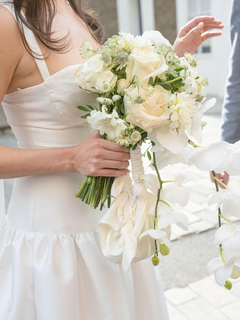 Bride holding a bouquet of white flowers and an ivory knotted handbag with a blurred background