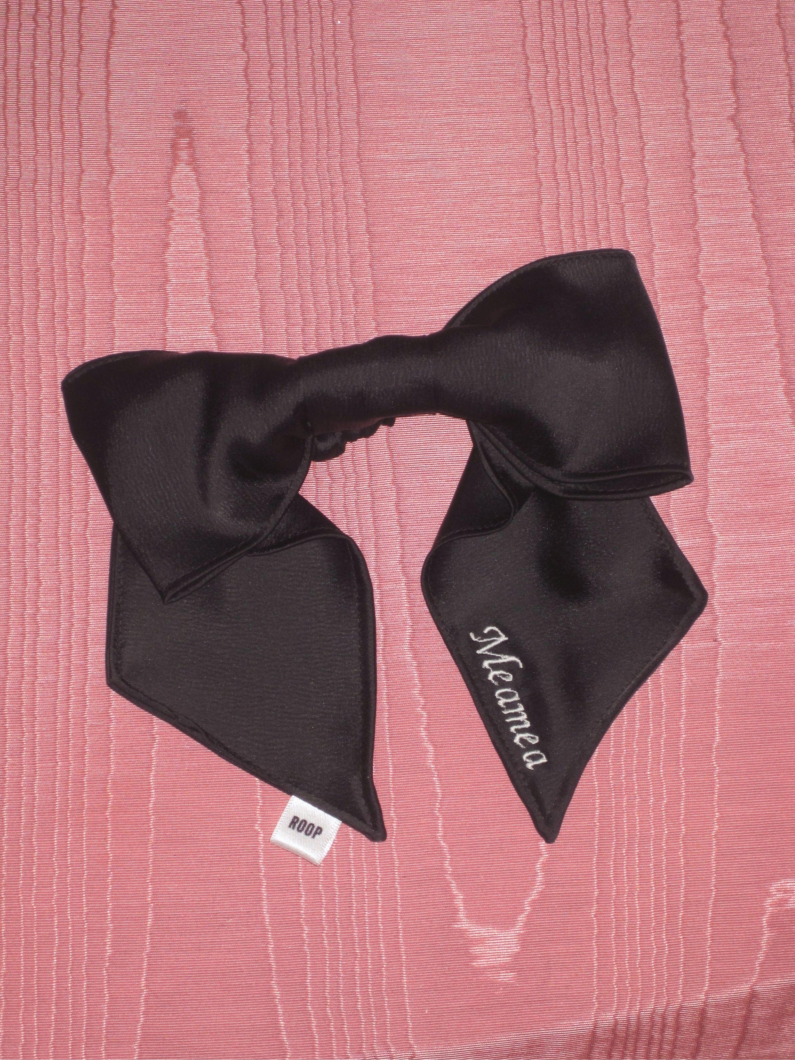 A black satin bow scrunchie with pale pink embroidery saying 'Meamea' on the tail. It is sat on a pink moire fabric.