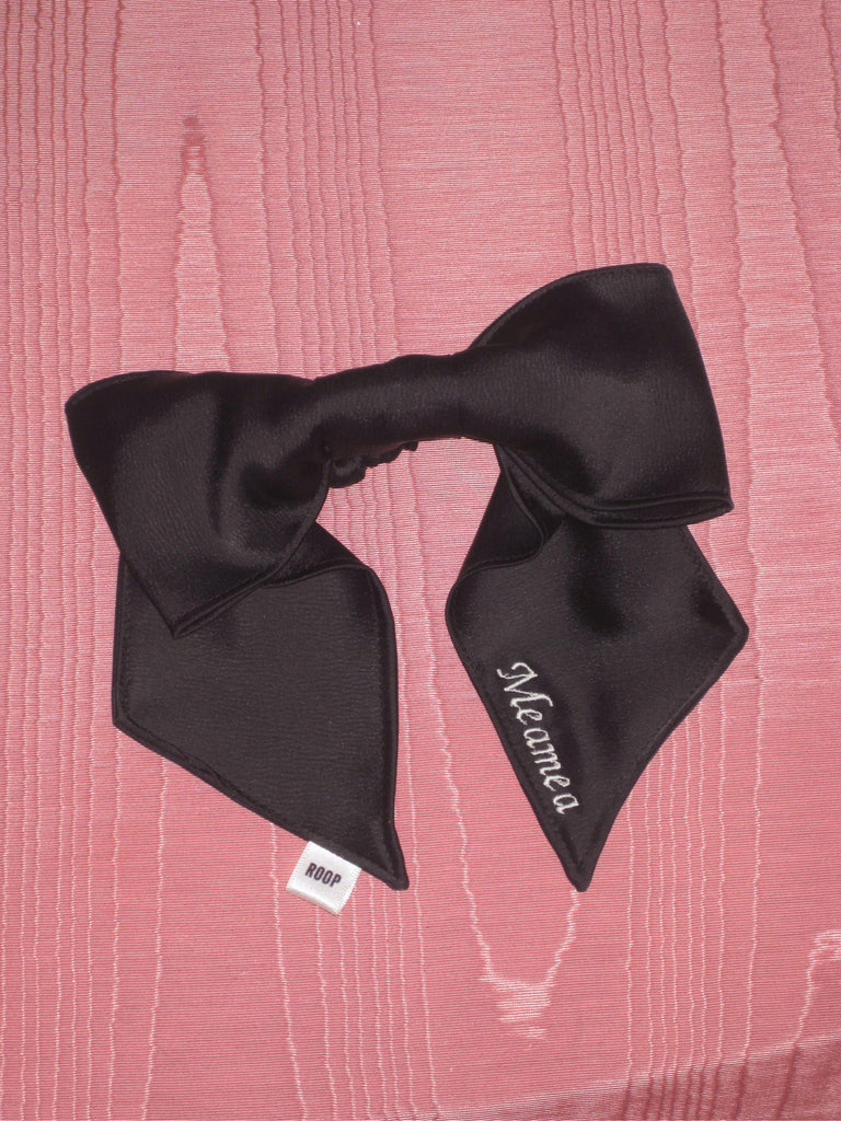 A black satin bow scrunchie with pale pink embroidery saying 'Meamea' on the tail. It is sat on a pink moire fabric.