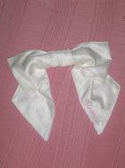 An ivory bamboo silk satin bow scrunchie with pale pink embroidery saying 'Meamea' on the tail. It is sat on a pink moire fabric.