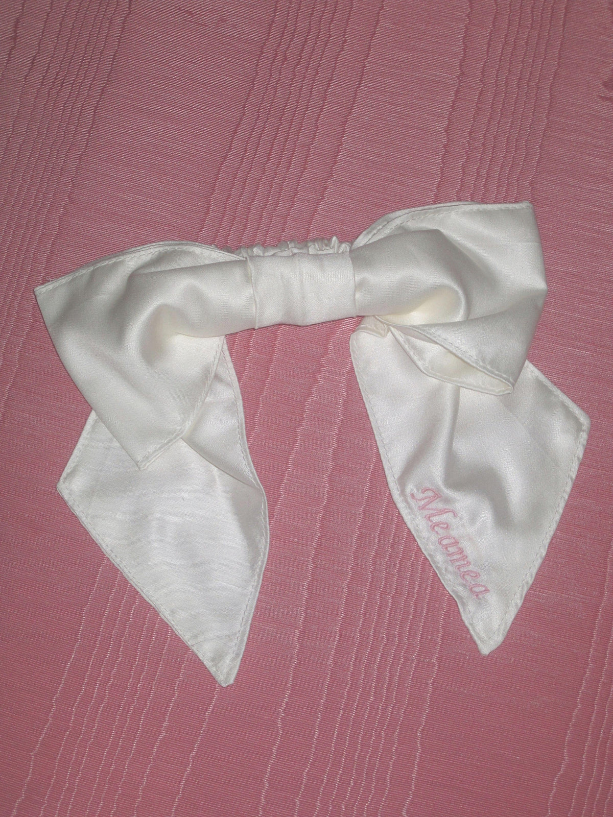 An ivory bamboo silk satin bow scrunchie with pale pink embroidery saying 'Meamea' on the tail. It is sat on a pink moire fabric.
