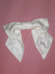 An ivory bamboo silk satin bow scrunchie with pale pink embroidery saying 'Meamea' on the tail. It is sat on a pink moire fabric.