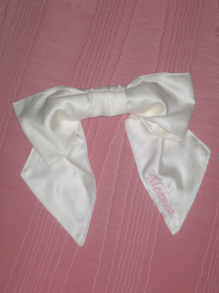 An ivory bamboo silk satin bow scrunchie with pale pink embroidery saying 'Meamea' on the tail. It is sat on a pink moire fabric.