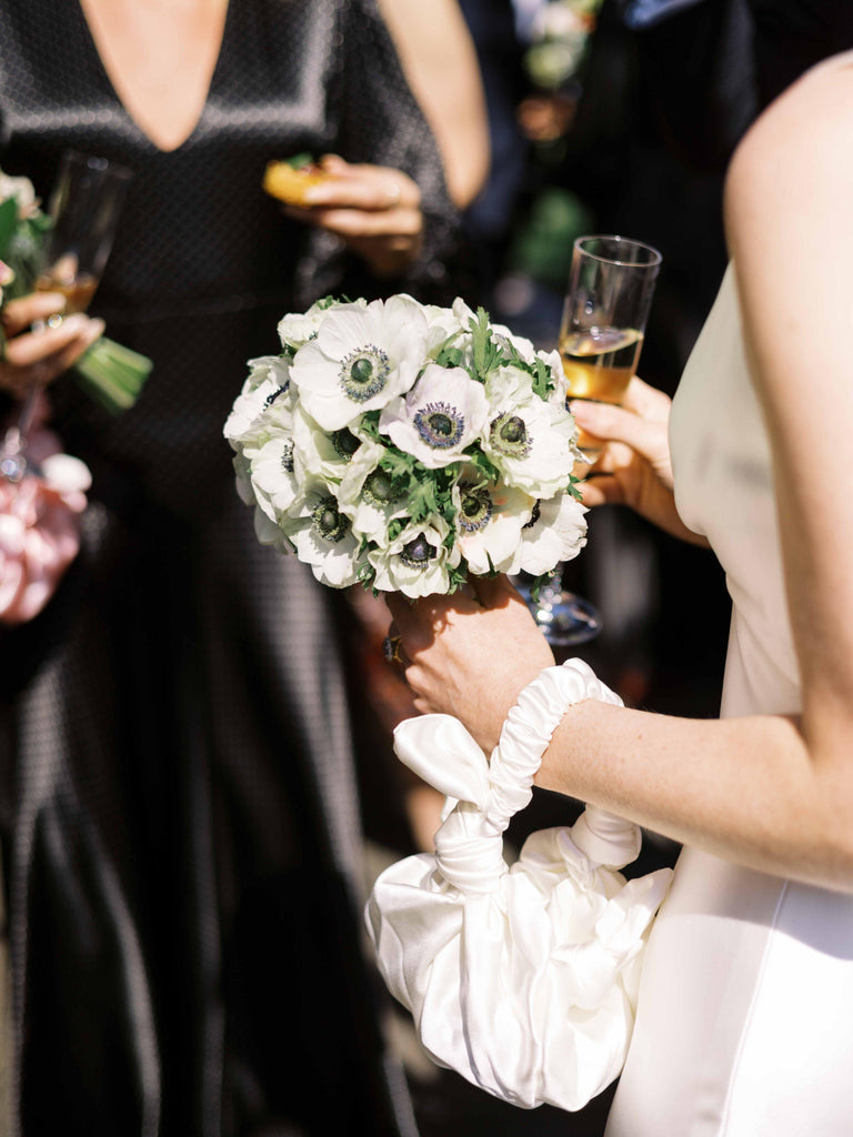 Bride holding a bouquet of white and green flowers with a glass of champagne and an ivory knotted handbag with a blurred background of people in black satin dresses.