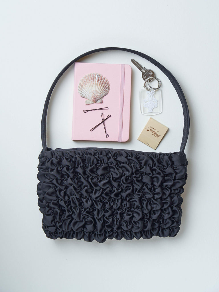 Black satin textured handbag with pink notebook, hairpins, and keychain on a light grey background.