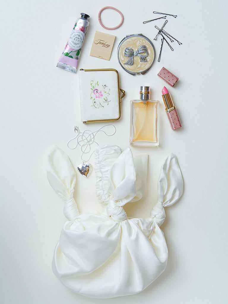 Collection of beauty products and accessories on a white surface, a white knotted handbag is at the bottom of the image.