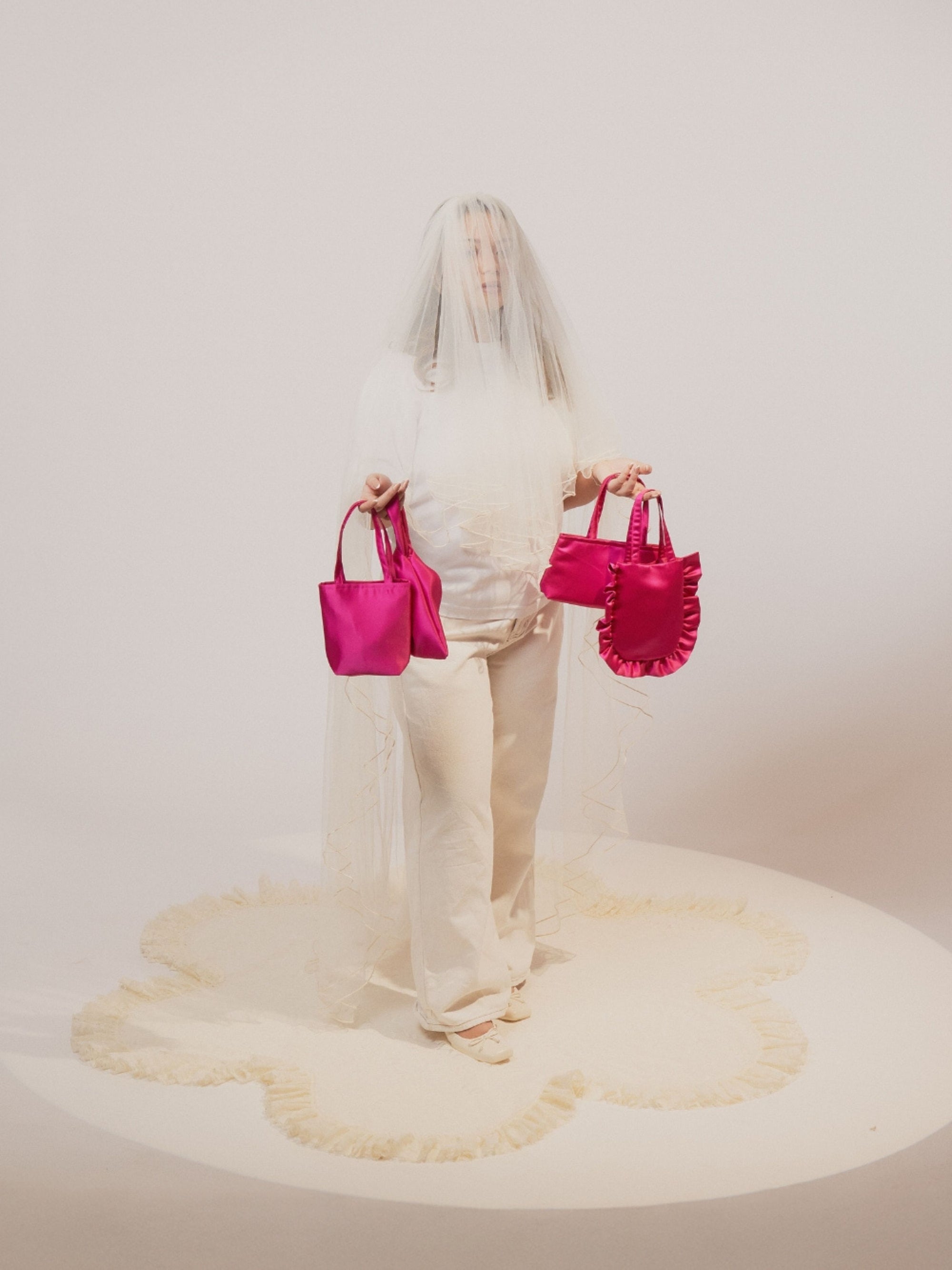 A person in a white outfit is partially veiled with sheer fabric, holding bold pink satin handbags. The scene emanates an artistic and surreal vibe.