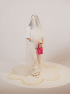 A person in a white outfit is partially veiled with sheer fabric, holding a bold pink satin handbag. The scene emanates an artistic and surreal vibe.