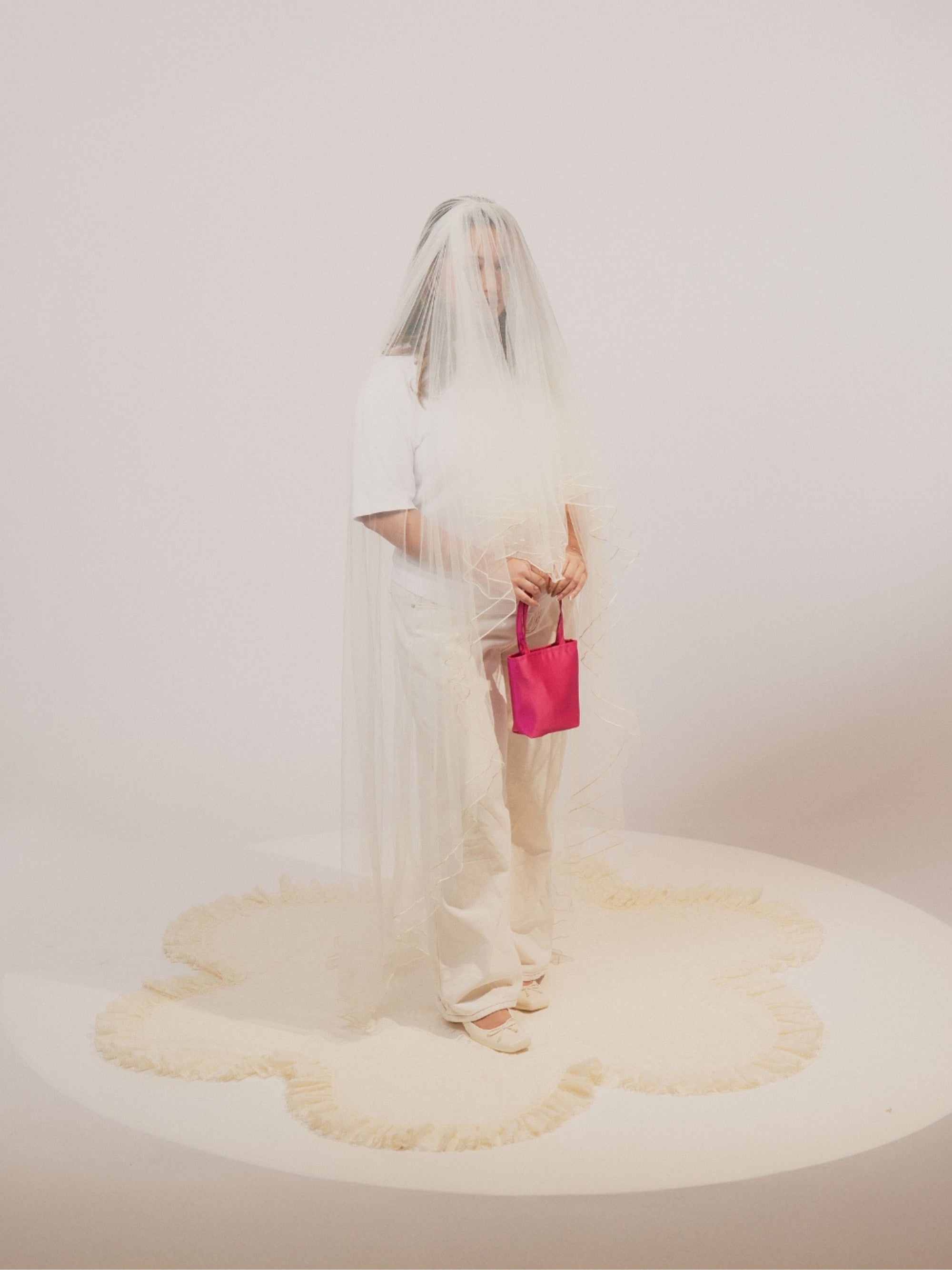 A person in a white outfit is partially veiled with sheer fabric, holding a bold pink satin handbag. The scene emanates an artistic and surreal vibe.