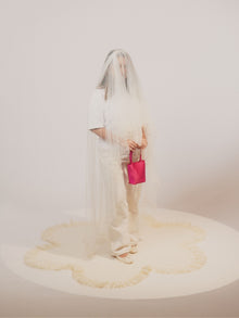 A person in a white outfit is partially veiled with sheer fabric, holding a bold pink satin handbag. The scene emanates an artistic and surreal vibe.