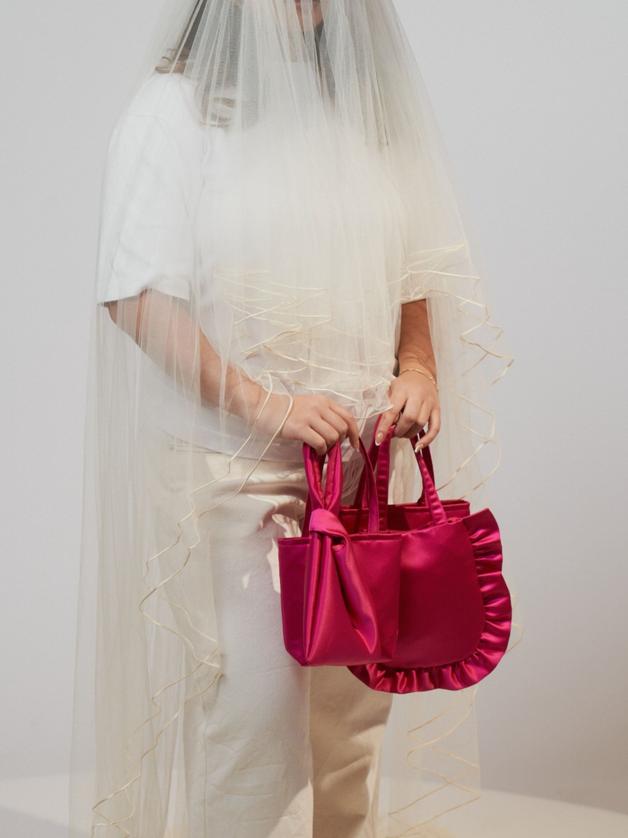 A person in a white outfit is partially veiled with sheer fabric, holding bold pink satin handbags. The scene emanates an artistic and surreal vibe.