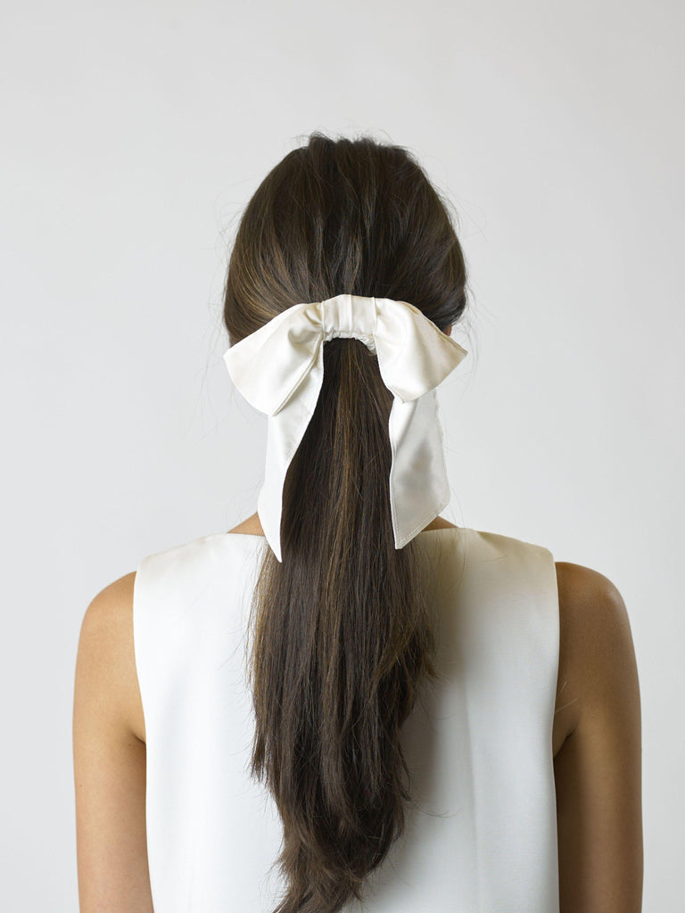 Person with long brown hair tied back with a large ivory bamboo silk satin hair bow scrunchie, against a plain background.